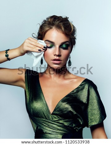 beautiful young girl with a bright make-up and in a shiny green dress striatet makeup from her face with a wet napkin. Hairstyle - curls are gathered in a bun.fashion, beauty, makeup, cosmetics.