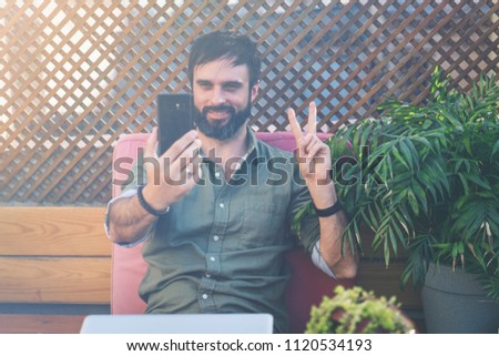 Content confident man in stylish clothes photographing himself on mobile phone and showing peace gesture.Bearded hipster making selfie via smartphone on terrace outside.