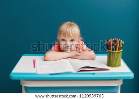 Portrait of little blonde girl is sitting at the table and paints. Schoolkid is doing her homework in classroom at school or at home. Tired and sad pupil