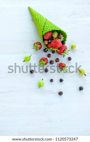 green ice cream cone with strawberries and currants and gooseberries, filled with strawberries it fell out of the horn, lies on a wooden white background, vegetarian food selective focus