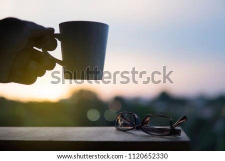 Hand holding a mug and eyewear glasses with sunset background.