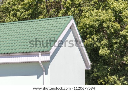 house with green tiled metal roof and white rain gutter on summer trees background