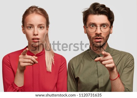 Emotive young female and male with amazed expressions, indicate at camera with index fingers, choose something, have scared looks, stand against white concrete wall. People and reaction concept
