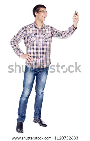 Full body portrait of smiling young man taking selfie on mobile phone wearing metal frame glasses, checkered shirt, blue jeans and black shoes isolated on white background