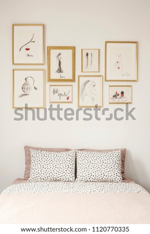 Stylish drawings gallery in golden frames above a cozy double bed with polka dot sheets in a white apartment room interior of a beauty lover