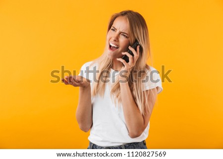Portrait of an angry young blonde girl talking on mobile phone isolated over yellow background