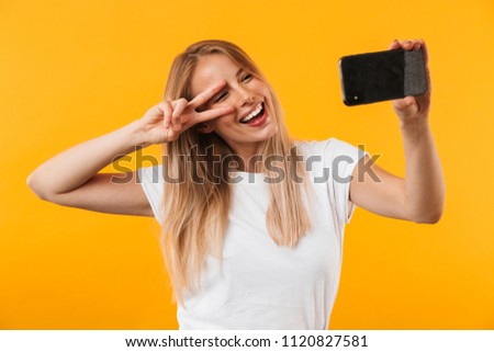 Lovely young blonde girl showing peace gesture while taking a selfie with mobile phone isolated over yellow background