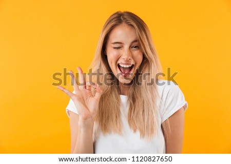 Portrait of a happy young blonde girl showing ok gesture and winking isolated over yellow background