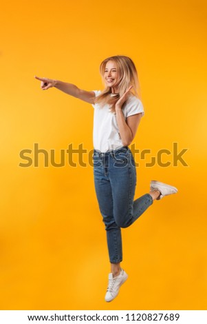Full length portrait of a satisfied young blonde girl pointing away while jumping isolated over yellow background