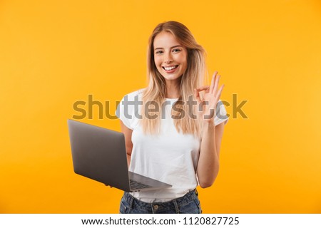 Portrait of a smiling young blonde girl holding laptop computer and showing ok gesture isolated over yellow background
