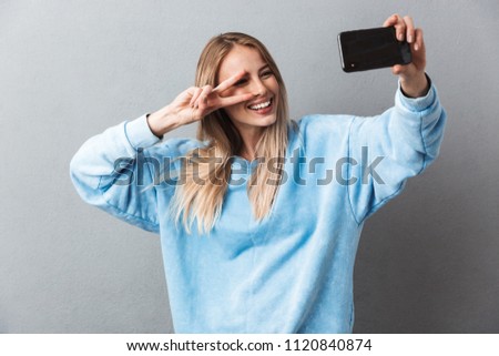 Smiling young blonde girl taking a selfie and showing peace gesture isolated over gray background