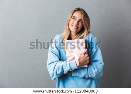 Portrait of a smiling pensive young blonde girl hugging present box isolated over gray background