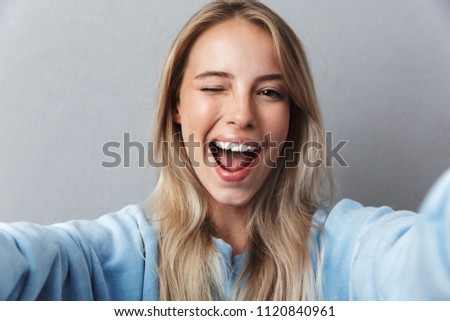Close up of smiling young blonde girl taking a selfie and winking isolated over gray background