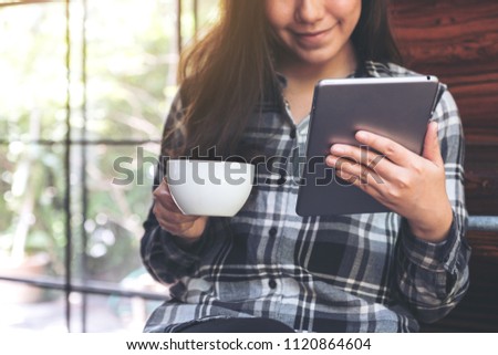 Closeup image of an asian woman holding and using tablet pc while drinking coffee in cafe