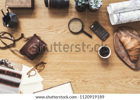 Stylish composition of flat lay on wooden desk with map, cup of coffee, croissant, phone, notebook, camera and office accessories. Creative desk of freelancer. Copy space for inscription.