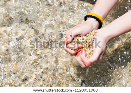 Beach sand in female hands by the sea, tiny underwater stones, sea bottom in background, tanned skin, sunlight daytime outdoors, summer vacation concept  