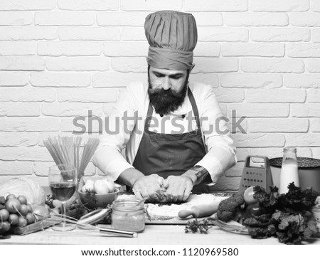 Cooking process concept. Chef makes dough. Man with beard kneads dough on white brick wall background. Cook with concentrated face in burgundy uniform sits by table with vegetables and kitchenware