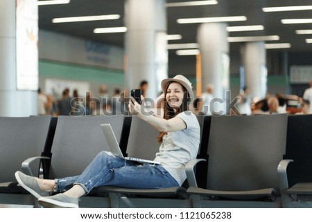 Young smiling traveler tourist woman working on laptop, doing selfie on mobile phone waiting in lobby hall at international airport. Passenger traveling abroad on weekends getaway. Air flight concept