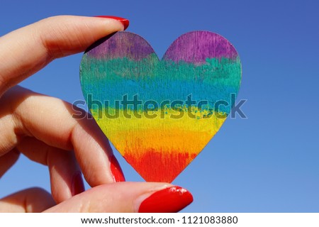 Female hand holding a rainbow colored wooden heart with a blue sky. LGBT / gay pride background concept.