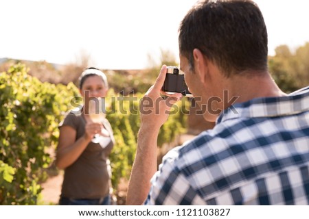 Man taking a photo of woman in a vineyard drinking a glass of wine and holding it toward the camera
