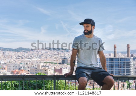Bearded muscular hipster man model wearing gray blank t-shirt and a black baseball cap with space for your logo or design in casual urban style.Green palm and cactus garden on the background
