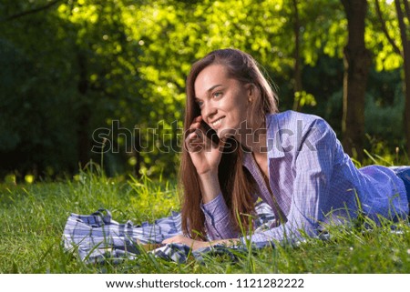 A girl in a shirt is lying on a plaid and talking on the phone in the park.