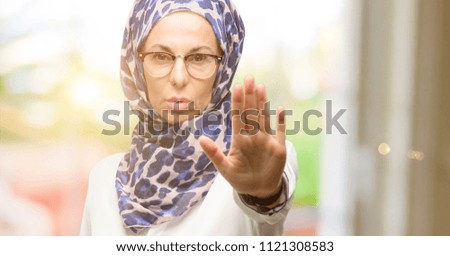 Middle age muslim arab woman wearing hijab annoyed with bad attitude making stop sign with hand, saying no, expressing security, defense or restriction, maybe pushing