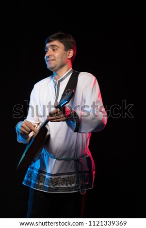 Artist musician A brunette man in a folk shirt playing a balalaika in scenic blue and red light on a black stage