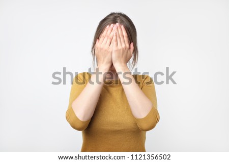 Young european woman in yellow sweater hides her face, studio photo isolated on gray background. She has social phobia, tying to be anonym Royalty-Free Stock Photo #1121356502