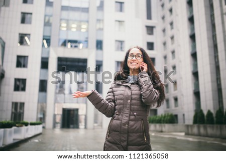 Theme is the business situation. Beautiful young woman of European ethnicity with long brunette hair wearing glasses and coat stands on background of business center and uses phone in hand near ear.
