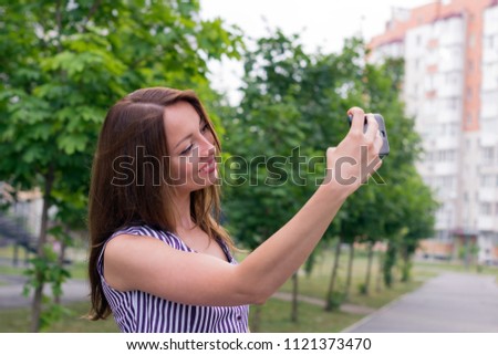 Portrait of young happy woman taking selfie in city smiling and posing for photo. Long-haired girl with nice appearance and sweet smile. Blurred background