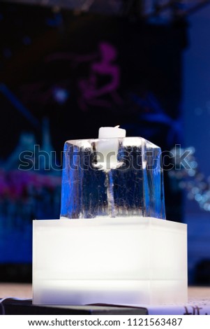 An ice sculpture with two ice blocks and a candle in the sculpture on a table with a white material with black letters with a blurry background .