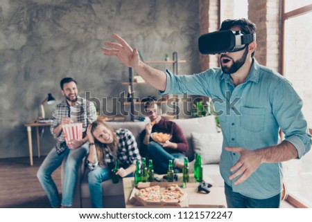 Portrait of amazed attractive bearded guy in vr-headset watching video trying to touch something with arms, his happy friends eating chips, popcorn on blurred background laughing at him