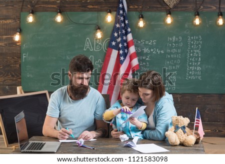 American family at desk with son making paper planes. Homeschooling concept. Parents teaching son to create, handmade. Kid with parents in classroom with usa flag, chalkboard on background.