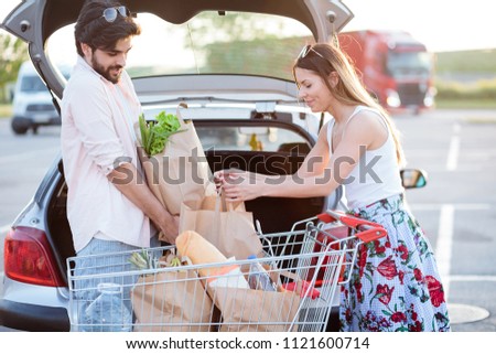 Happy young couple loading grocery bags into a car trunk at a parking lot in front of a shopping mall. Boyfriend lifts heavy bag while girlfriend is helping. Royalty-Free Stock Photo #1121600714