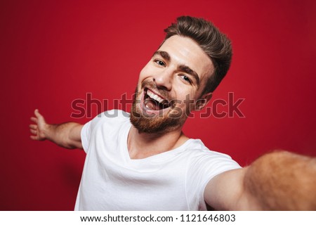 Portrait of a smiling young bearded man taking a selfie isolated over red background