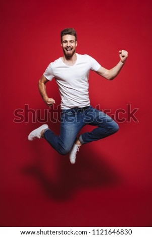 Full length portrait of a cheerful young bearded man celebrating success while jumping isolated over red background