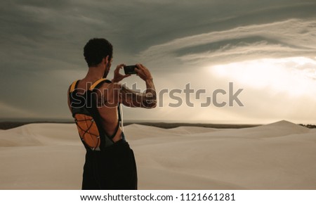 Rear view of man taking pictures of dramatic sky with his mobile phone during fitness training in desert. Fit man capturing cloudy sky pictures in his mobile phone while workout break.