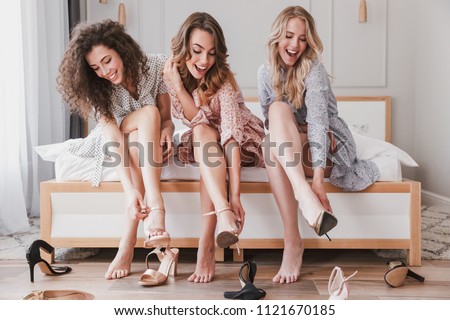 Picture of three gorgeous girls 20s wearing dresses trying on different summer stilettos or high heels during bachelorette party in posh bedroom