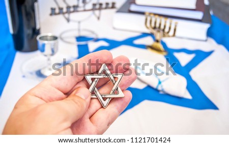 Hand holding a David Star ("Magen David" in Hebrew), a traditional Jewish religious symbol. With more Jewish symbols on the background: prayer books, menorah lampstand, kiddush wine blessing glass.