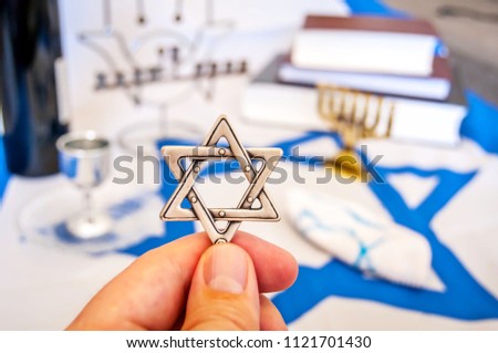 Hand holding a David Star ("Magen David" in Hebrew), a traditional Jewish religious symbol. Prayer books, menorah lampstand, kiddush wine blessing glass. Conversion to Judaism, giyur.