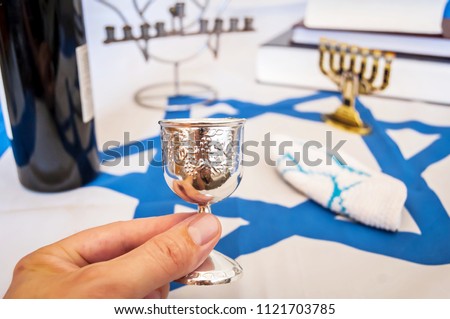 Hand holding a "kiddush" wine blessing glass, a traditional Jewish religious symbol. With more Jewish symbols on the background: prayer books, "menorah" lampstand on a flag of Israel.