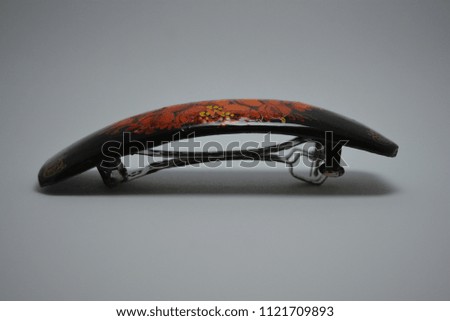 On a white paper background lies and is a black wooden barrette with a metal clip for hair, painted with black paint with red flowers painted on it