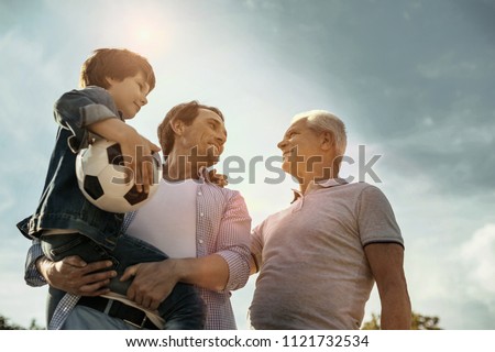 Feeling proud. Exuberant smiling loving daddy son and grandfather looking at each other while the dad holing his son in his hands and the son holding his ball Royalty-Free Stock Photo #1121732534