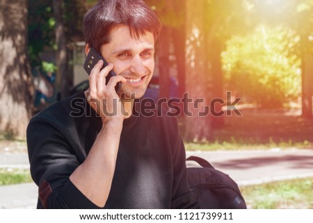 A young man with black hair and sweaters smiles and talks on a smartphone on a nature background, Toned, close-up