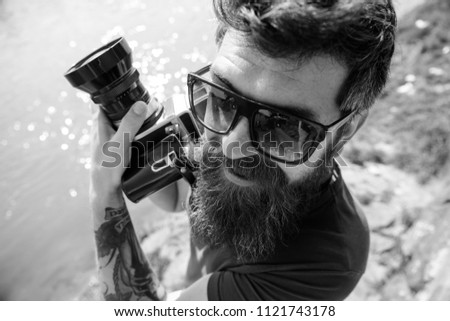 Tourist photographer concept. Man with beard and mustache wears sunglasses, water surface on background. Guy shooting nature near river or pond. Hipster on smiling face holds old fashioned camera.