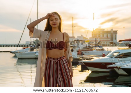 slender luxury girl in stylish summer pants is waiting for her yacht in the port in the evening, smiling