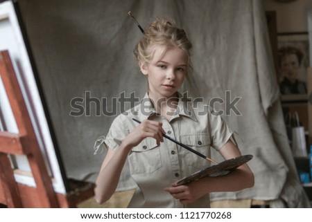 Little artist girl holding a paintbrush and looking over a canvas on an easel. Concept of creativity and fun