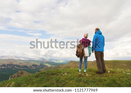 A pair of hipster tourists is standing next to each other in the mountains against the backdrop of the plateau of valleys and sky. Rest in the mountains. Traveling in pairs