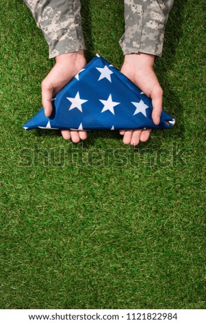 partial view of soldier in military uniform holding folded flag in hands on green grass, 4th july holiday concept